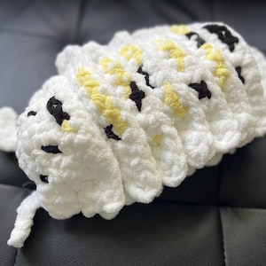 Puede incluir: Un juguete de peluche blanco tejido a crochet con detalles amarillos y negros. El juguete tiene forma de oruga y tiene un cuerpo segmentado.