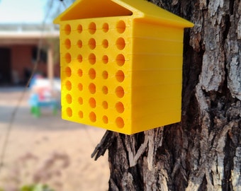 May include: A yellow bee house with a roof and multiple holes for bees to nest in. The bee house is attached to a tree trunk.