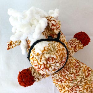 May include: A handmade crochet horse toy with a white mane and a black bridle. The body is a mix of cream, orange, and brown yarn, with red accents on the hooves. This soft toy is perfect for children.