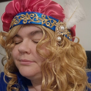 May include: A pink satin cap with a blue band and gold embroidery. A gold brooch with pearls and a white feather are attached. This headwear is a costume piece, possibly for a play or themed occasion. The design is reminiscent of historical styles.