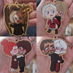 May include: A set of four wooden keychains featuring colourful chibi-style illustrations of fantasy characters. The designs include a character with a dragon, two figures in red and black, a couple kissing, and two characters embracing in a hug.