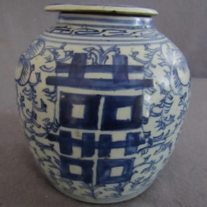 May include: A blue and white ceramic jar with a lid. The jar features a large, stylized double happiness symbol in the center, surrounded by floral and leaf patterns. The jar is a traditional Asian design.