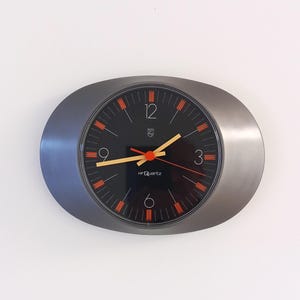 Op de afbeelding: Een ovale wandklok met een donkergrijze metalen rand en een zwarte ronde wijzerplaat. De wijzerplaat heeft witte cijfers op 12, 3, 6 en 9, oranje uurmarkeringen, gele uren- en minutenwijzers en een rode secondewijzer. De tekst HF Quartz is zichtbaar.