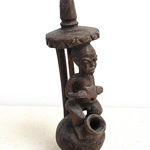 Op de afbeelding: Een gesneden houten sculptuur met een zittende figuur op een bolvormige basis met een tuit. De figuur bevindt zich onder een decoratieve paraplu-achtige structuur. De sculptuur is donkerbruin van kleur en heeft een rustieke uitstraling.