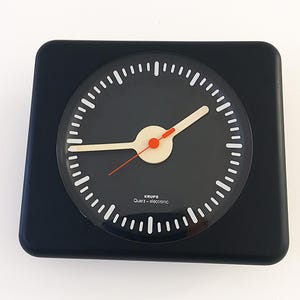 Op de afbeelding: Een vierkante zwarte wandklok met een ronde zwarte wijzerplaat, witte streepjes en lichtgekleurde uren- en minutenwijzers. Een dunne rode secondewijzer komt uit het midden. De tekst KRUPS Quarz-electronic is in het wit op de wijzerplaat gedrukt.