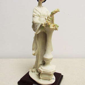 Può includere: Statuetta color crema di una donna in abito asiatico tradizionale, con un vaso di fiori. La statuetta è montata su una base rettangolare marrone scuro. La scultura è alta circa 25 cm.