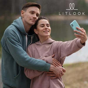 May include: A couple wearing matching hoodies, one teal and the other dusty rose, pose for a selfie. The woman holds a teal smartphone, smiling. The hoodie features a hood and a front pocket. The brand name "LITLOOK" is displayed in the upper right corner.