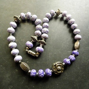 May include: A purple and silver beaded necklace with a floral design. The necklace is made of ceramic beads with a white floral pattern and silver metal beads.
