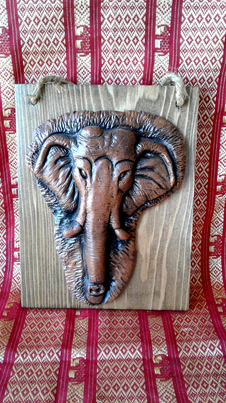 May include: A copper-coloured elephant head sculpture mounted on a rectangular wooden plaque. The elephant has detailed features, including large ears and a long trunk. A natural rope is attached for hanging. The background is a red and gold patterned fabric.
