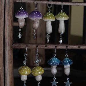 May include: A selection of handmade mushroom earrings in shades of purple, green, yellow, and blue. Each earring showcases a distinctive mushroom cap design with white polka dots, a beige stem, and a dangling silver charm. The earrings are presented on a wooden rack.
