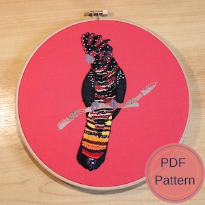 May include: Embroidered hoop art featuring a black and red bird with yellow and orange accents perched on a branch. The background is a solid red colour. The text "PDF Pattern" is visible in the lower right corner.