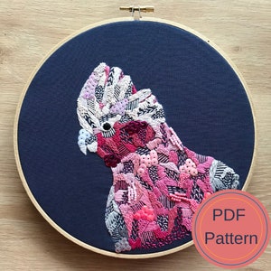 May include: Embroidered pink and white cockatoo on a dark blue fabric background in a wooden embroidery hoop. The text "PDF Pattern" is in a pink circle in the lower right corner of the image.