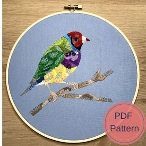 Può includere: Un uccello colorato ricamato su un ramo, con uno sfondo blu. L'uccello è un fringuello con la testa rossa e il corpo giallo, verde e viola. Il ramo è marrone e ha alcune foglie. Il ricamo è in un cerchio, e lo sfondo è un tessuto blu chiaro. L'immagine include il testo "PDF Pattern".