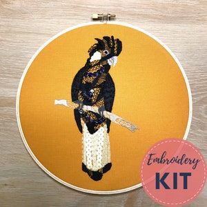 May include: An embroidery kit featuring a black cockatoo perched on a branch, set against a mustard-colored fabric within a wooden hoop. The bird is detailed with black, gold, and white thread, and embellished with blue sequins. The text "Embroidery KIT" is in a pink circle.