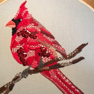 May include: Close-up of a red cardinal embroidered with sequins and beads, perched on a brown branch.
