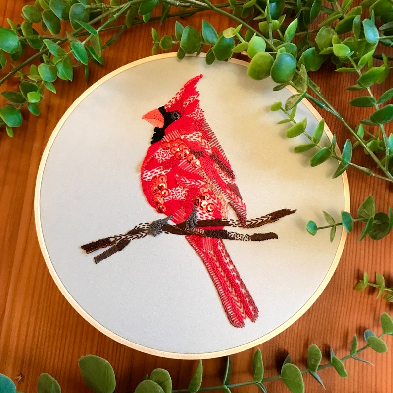 May include: Embroidered cardinal bird on a light blue background. The cardinal is red with black eyes and a black beak. The bird is perched on a brown branch.