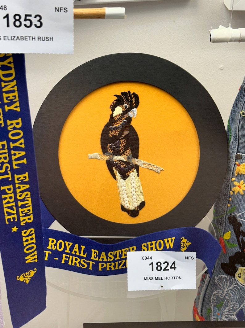 May include: A framed embroidery of a black cockatoo perched on a branch, set against a yellow background. The bird is detailed with various thread colors and textures. The frame is black and round. A blue ribbon with gold text and a white tag are also visible.