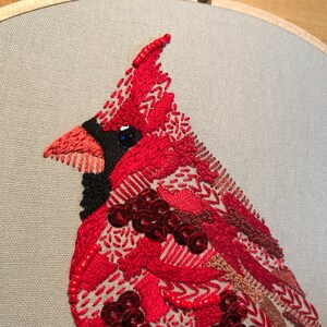 May include: Close-up of a red cardinal embroidered on a light gray fabric. The cardinal is made with red, black, and brown thread, and has a blue eye. The embroidery is detailed and intricate, with sequins and beads used to add texture.