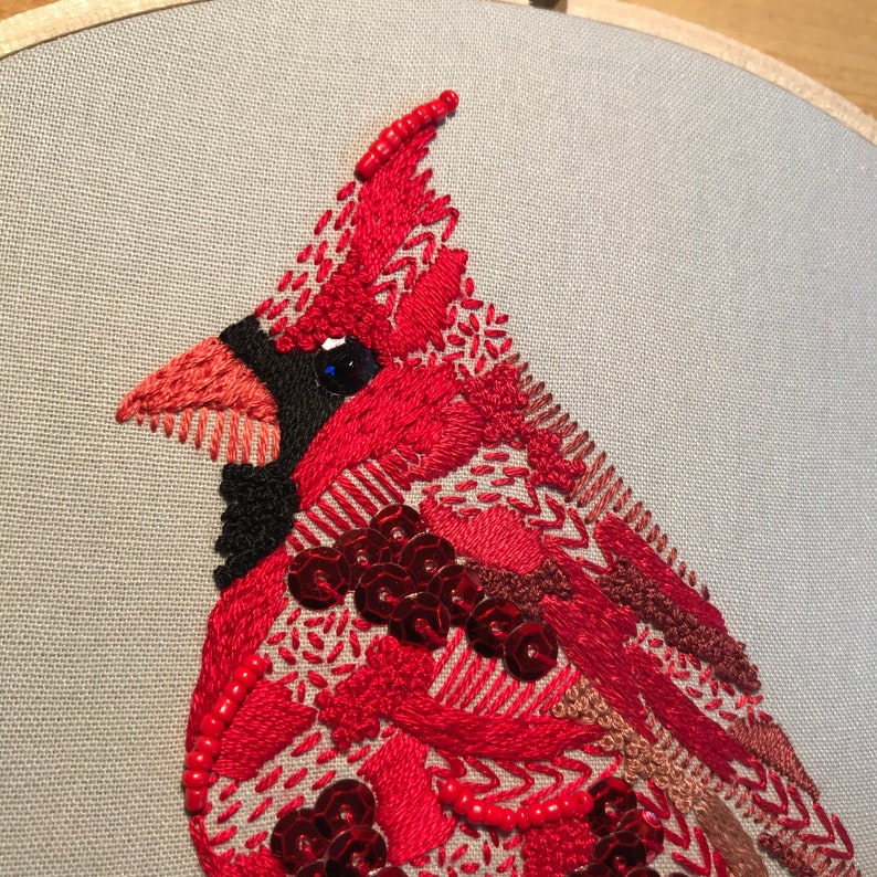 May include: Close-up of a red cardinal embroidered on a light gray fabric. The cardinal is made with red, black, and brown thread, and has a blue eye. The embroidery is detailed and intricate, with sequins and beads used to add texture.