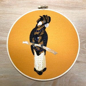 May include: Embroidered black and white bird with a long tail, perched on a branch, on a yellow background. The bird is surrounded by a brown wooden hoop.