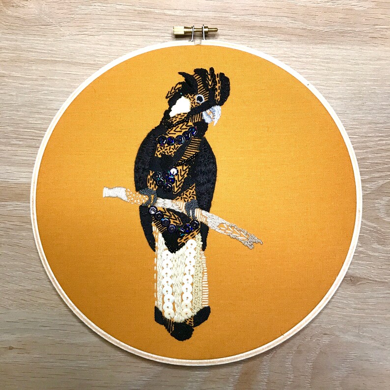 May include: Embroidered black and white bird with a long tail, perched on a branch, on a yellow background. The bird is surrounded by a brown wooden hoop.