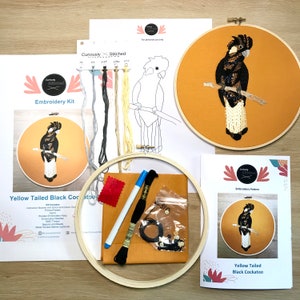 May include: An embroidery kit featuring a yellow-tailed black cockatoo design. The kit includes a wooden embroidery hoop, embroidery needles, DMC thread, a printed pattern, and a water-soluble marker. The kit also includes a color guide and instructions.