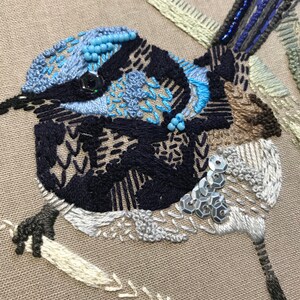 May include: Close-up of an embroidered bluebird with black, white, and brown thread. The bird is decorated with blue and silver beads and sequins.