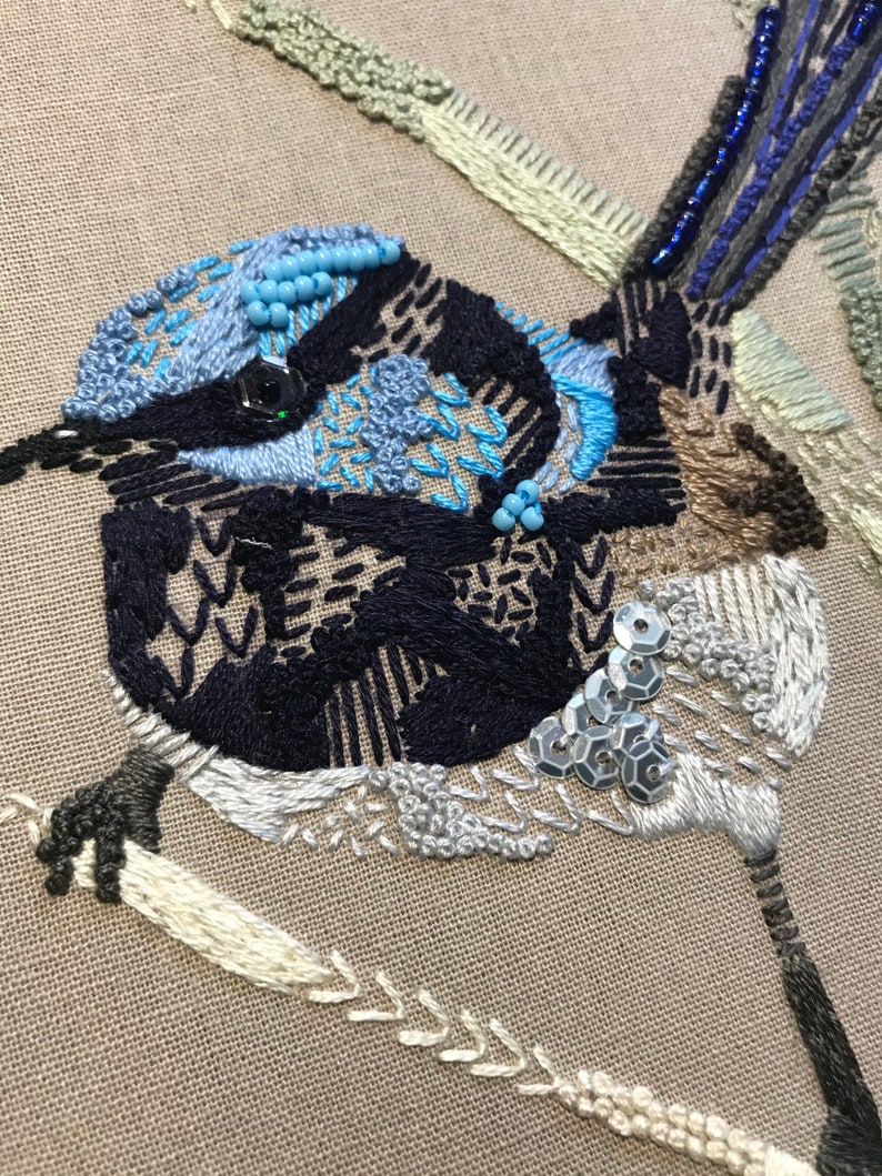 May include: Close-up of an embroidered bluebird with black, white, and brown thread. The bird is decorated with blue and silver beads and sequins.