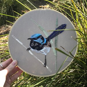May include: Embroidered artwork of a blue and black bird with a long tail, perched on a branch with green and white leaves. The artwork is framed in a wooden hoop.