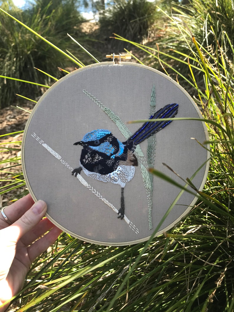 May include: Embroidered artwork of a blue and black bird with a long tail, perched on a branch with green and white leaves. The artwork is framed in a wooden hoop.