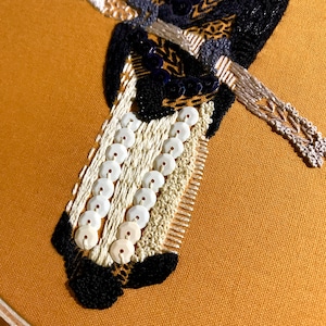 May include: Close-up of an embroidered artwork on a mustard-colored fabric. The embroidery features black, white, and silver threads, along with white sequins. The design includes geometric patterns and detailed stitching, creating a textured and ornate appearance.