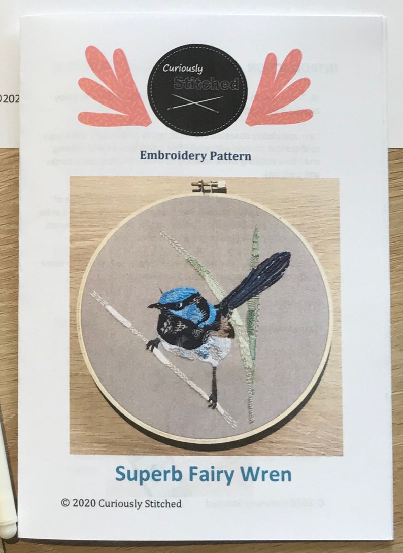 May include: Embroidery pattern for a Superb Fairy Wren, a small blue bird with a black head and white belly, perched on a branch with green leaves. The pattern is from Curiously Stitched and is copyrighted 2020.