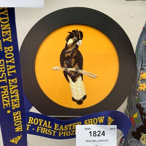 May include: An embroidered cockatoo in black and white on a yellow background, framed in black. The embroidery is detailed and includes white beads for the cockatoo's chest. The frame is surrounded by a blue ribbon with the text "Sydney Royal Easter Show - First Prize" and "Royal Easter Show - First Prize".