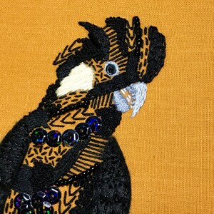 May include: Close-up of a black and white embroidered bird with a white beak and black eyes. The bird is on a brown fabric background.