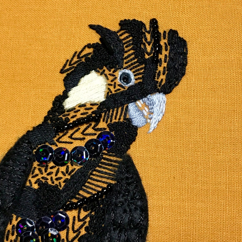 May include: Close-up of a black and white embroidered bird with a white beak and black eyes. The bird is on a brown fabric background.