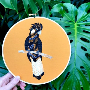 May include: A black and white embroidered bird with a yellow beak on a mustard yellow background. The bird is perched on a branch with black and white details. The embroidery is framed in a round wooden hoop.