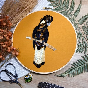 May include: Embroidered hoop art featuring a black and white cockatoo perched on a branch. The bird is surrounded by a mustard yellow background.