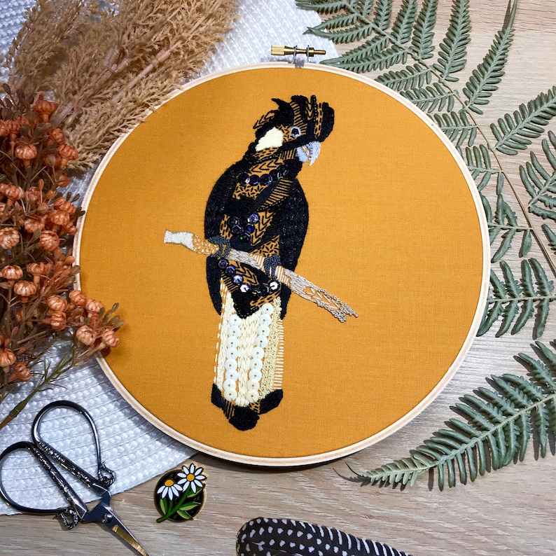 May include: Embroidered hoop art featuring a black and white cockatoo perched on a branch. The bird is surrounded by a mustard yellow background.
