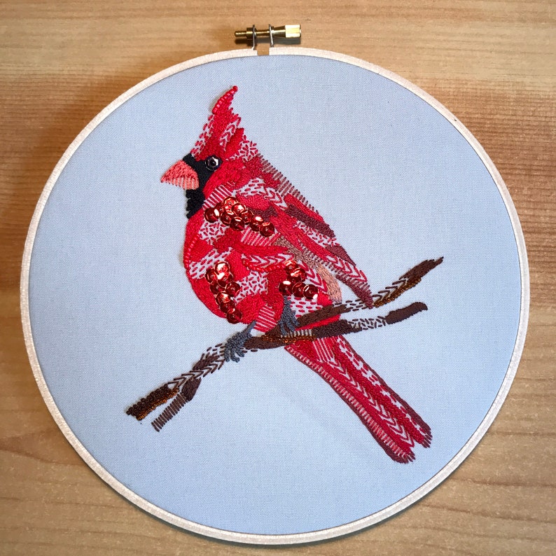 May include: Embroidered cardinal perched on a branch, with red, white, and brown thread details. The cardinal is on a light blue background, framed in a wooden hoop.