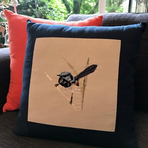 May include: A navy blue cushion with a white embroidered bird design. The bird is blue and black with a long tail and is perched on a branch with green leaves.