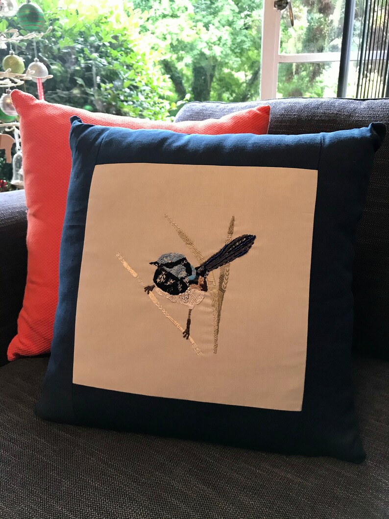 May include: A navy blue cushion with a white embroidered bird design. The bird is blue and black with a long tail and is perched on a branch with green leaves.