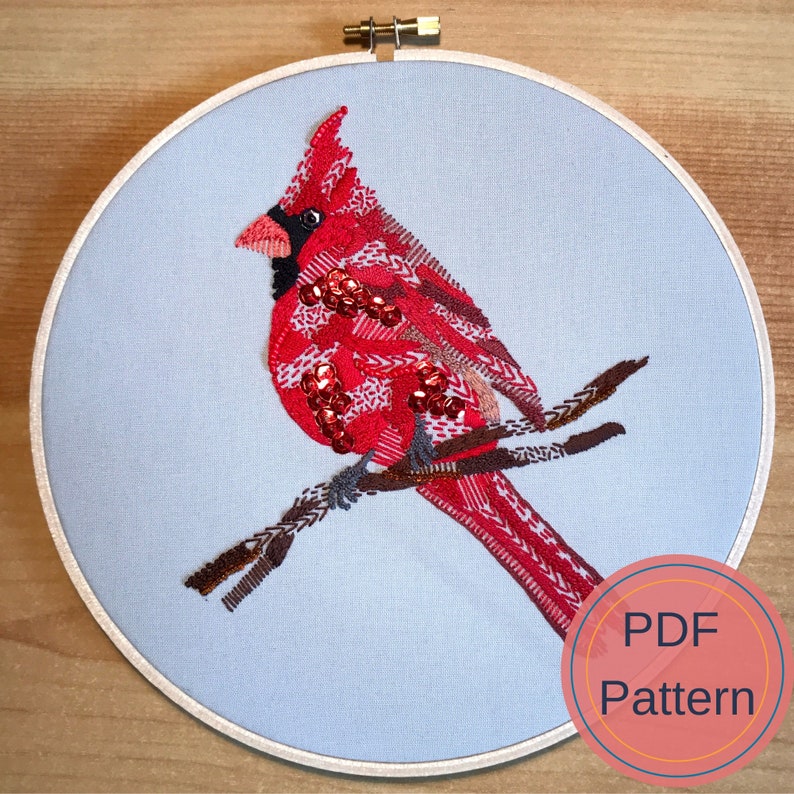 May include: Embroidered cardinal bird on a branch with red, brown, and white thread. The bird is on a light blue background in a round embroidery hoop. The text "PDF Pattern" is on a pink circle in the lower right corner of the image.