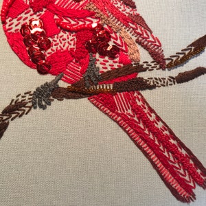May include: Close-up of a red embroidered bird with sequins and beads perched on a brown branch. The bird's tail feathers are detailed with intricate stitching.