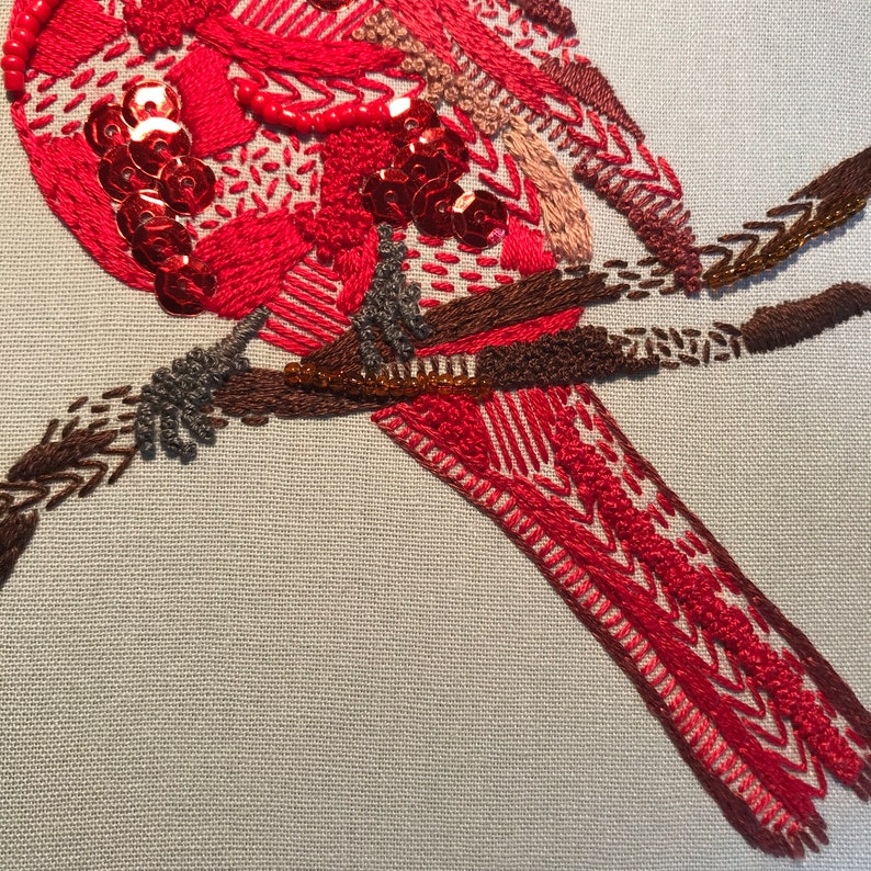 May include: Close-up of a red embroidered bird with sequins and beads perched on a brown branch. The bird's tail feathers are detailed with intricate stitching.