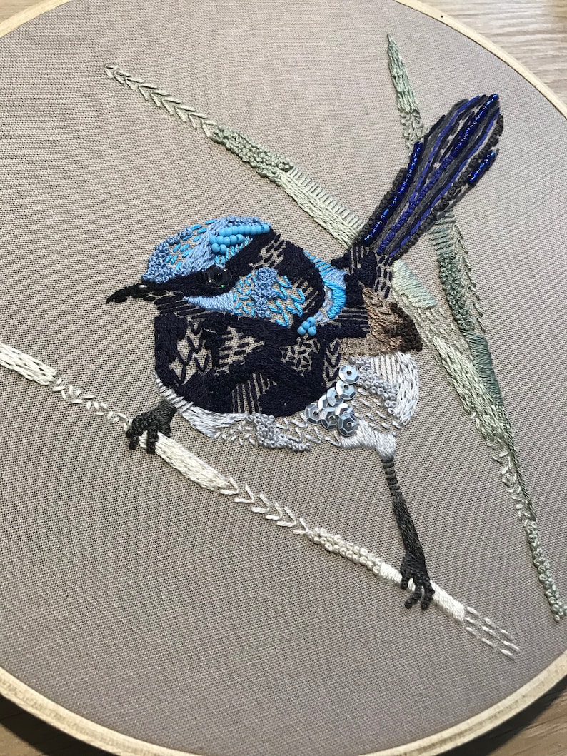 May include: Embroidered artwork depicting a blue and black bird perched on a branch. The bird has a long tail and is surrounded by green leaves.