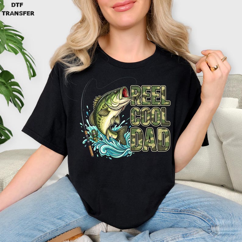 May include: Black t-shirt featuring a graphic of a leaping fish with water splashes and a fishing rod. The text "REEL COOL DAD" is in a camouflage pattern. The shirt is a DTF transfer.