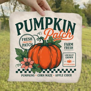 May include: A cream-colored fabric square featuring a large orange pumpkin graphic with green leaves and a pink flower. Text includes PUMPKIN PATCH, FRESH FROM THE PATCH, FARM FRESH, LOCALLY GROWN, AUTUMN PICKED, PUMPKINS, CORN MAZE, and APPLE CIDER.