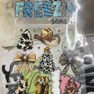 May include: Clear plastic sheet with various Christmas-themed graphics. Includes a cowboy hat, Christmas trees with cow print, bows, and cowboy boots. The text "It's Freezin' Season" is at the top.