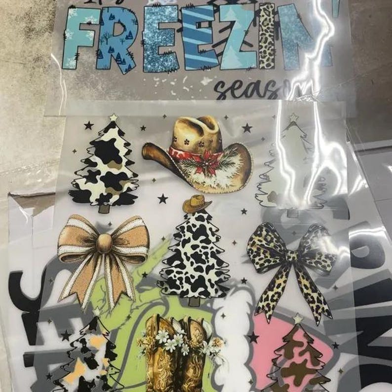 May include: Clear plastic sheet with various Christmas-themed graphics. Includes a cowboy hat, Christmas trees with cow print, bows, and cowboy boots. The text "It's Freezin' Season" is at the top.