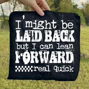 May include: Black square textile with white distressed text: "I might be LAID BACK but I can lean FORWARD real quick." The bottom has a black and white checkered pattern. Held up against a green grassy background.
